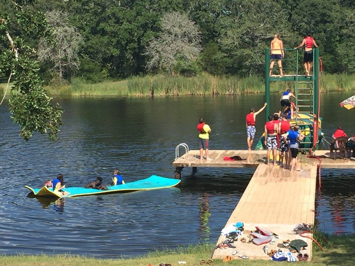 A lively scene by the private pond features guests engaging in outdoor activities. A dive tower is visible beside a wooden dock, with people climbing stairs and preparing to jump. A colorful kayak is seen on the water, while others gather on the dock.