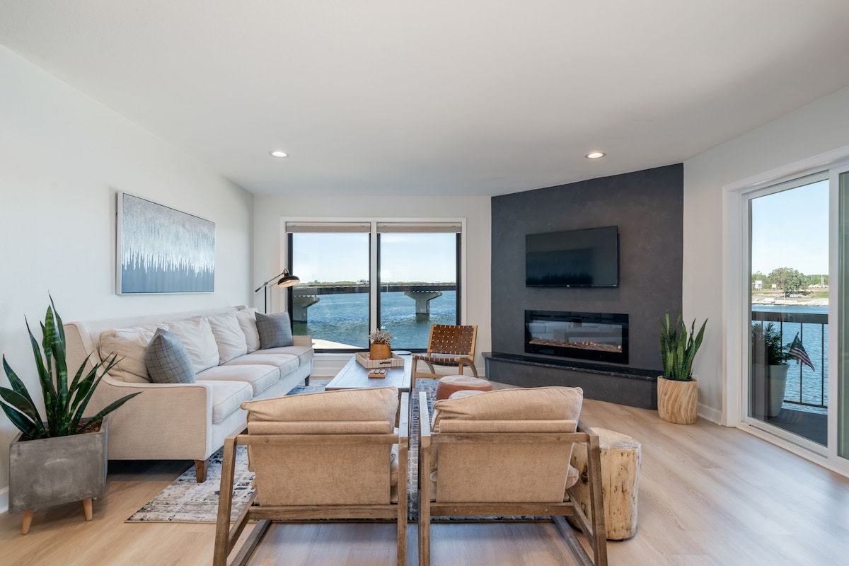 A modern living space is showcased with a light-colored sectional sofa and two armchairs positioned around a wooden coffee table. Large windows allow natural light and offer views of the lake. A wall-mounted television and a decorative fireplace enhance the ambiance.