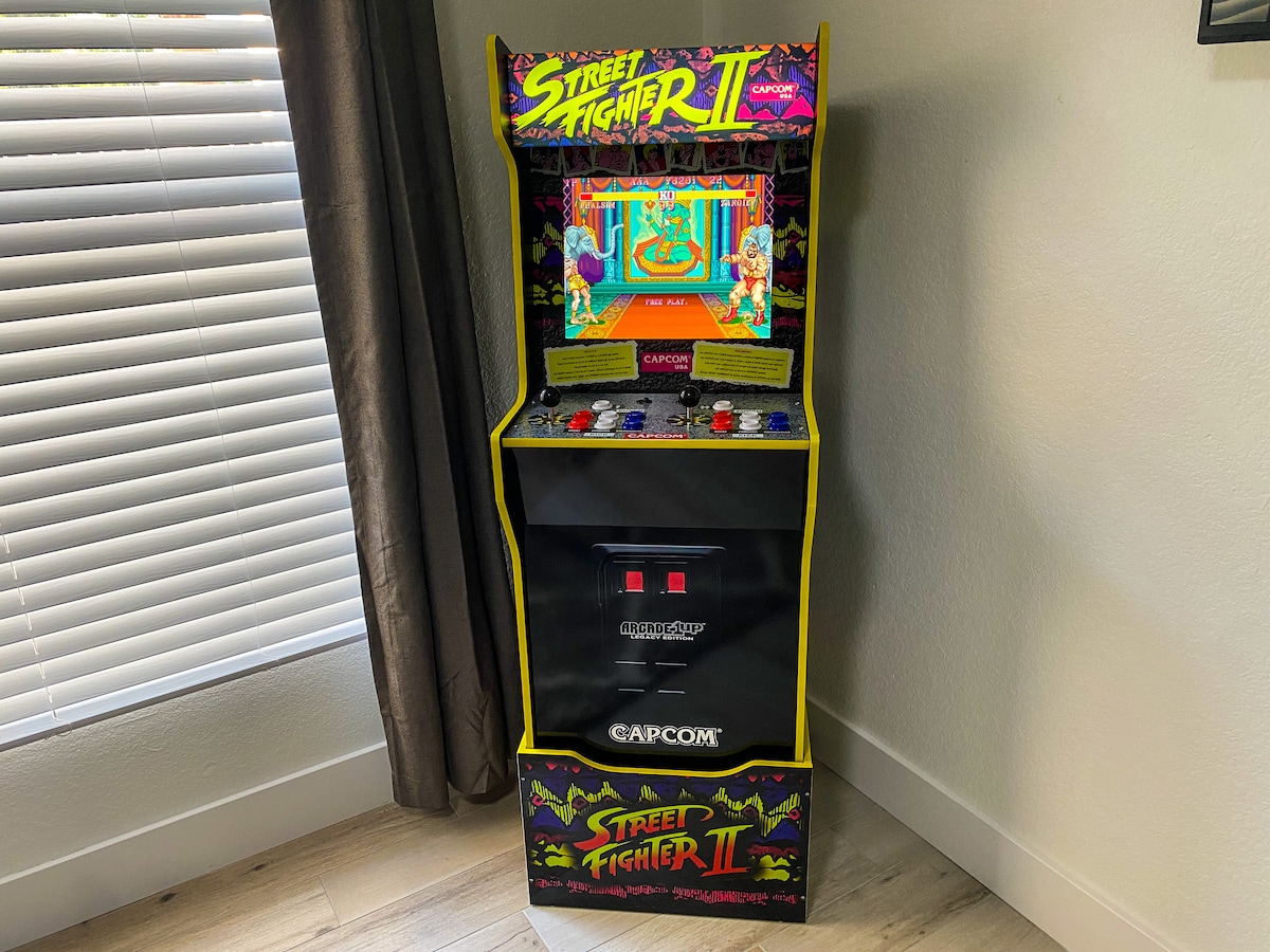 A stand-up arcade game featuring the classic 'Street Fighter II' title is placed against a wall. The brightly colored cabinet showcases arcade controls, including buttons and a joystick. Nearby, natural light filters through partially closed blinds, creating a well-lit and inviting gaming corner.