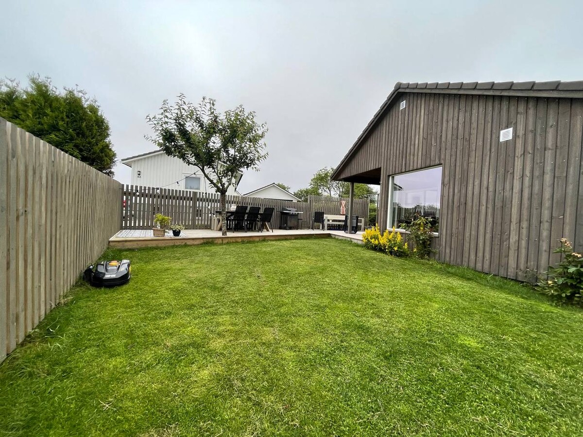 A well-maintained garden features a green lawn bordered by a wooden fence. A wooden terrace is visible, with a dining table set under an overcast sky. Surrounding plants add a touch of color, while the modern building offers large glass doors reflecting the outdoor space.