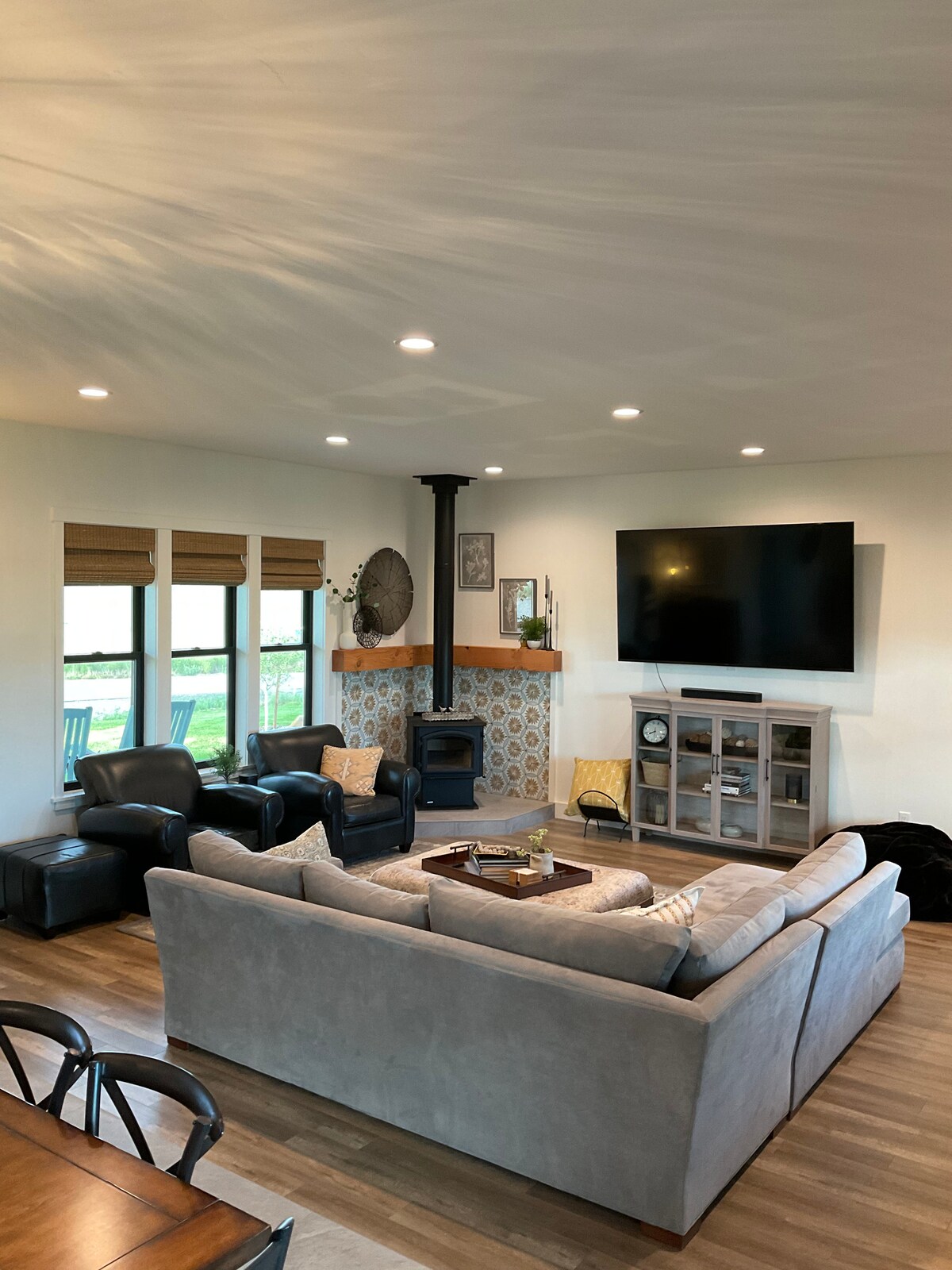 A spacious living area is highlighted by a comfortable sectional sofa and armchairs. Natural light enters through multiple windows, and a television is mounted on the wall opposite a modern gas stove. Decor includes patterned tiles and simple artwork.