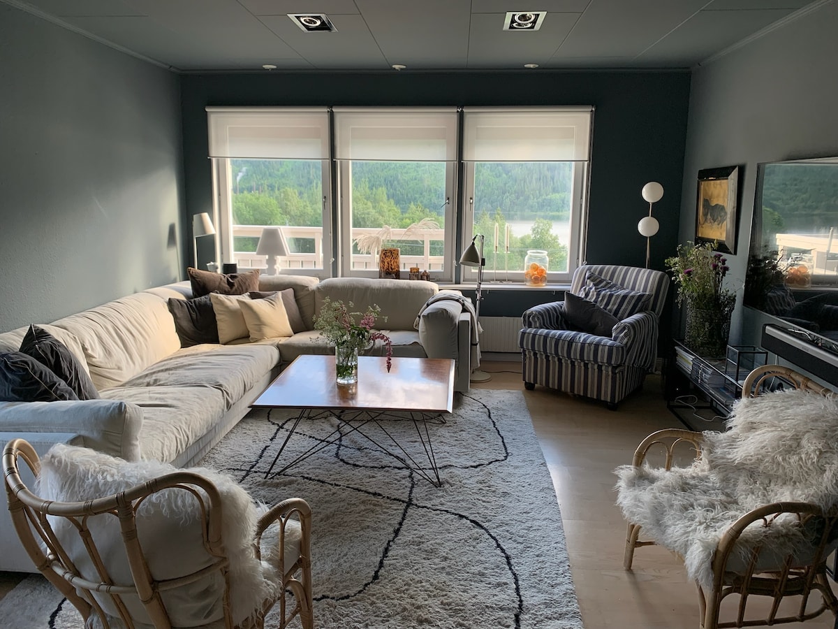 A light-filled living area features large windows revealing scenic views of the lake and mountains. A comfortable sofa is paired with a striped armchair, while a coffee table sits on a soft area rug. Decorative plants and tasteful accents enhance the relaxing space.