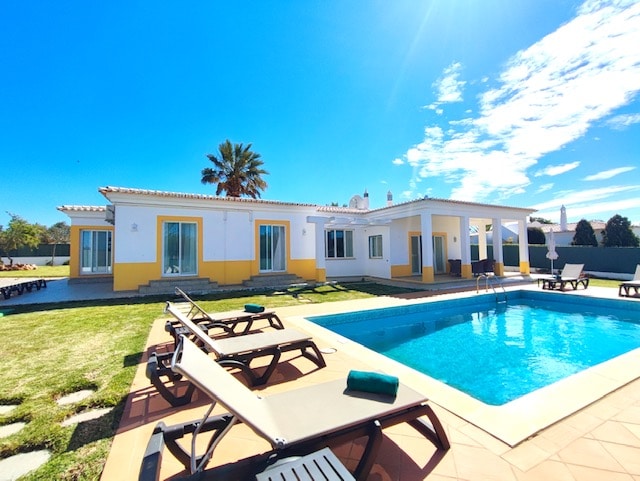 The exterior of a villa is displayed, featuring a clear blue sky and well-maintained gardens. A private pool is situated in front, surrounded by lounge chairs. The villa's architecture includes large windows and a covered patio area that offers shaded seating.