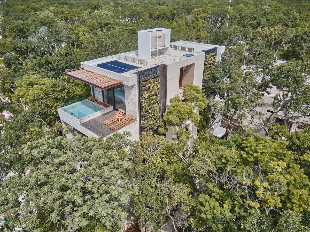 An aerial view captures a modern penthouse nestled among lush greenery. The building features sleek architectural lines and expansive terraces. A private jacuzzi is visible on the balcony, complemented by lounge chairs, while solar panels are situated on the rooftop.