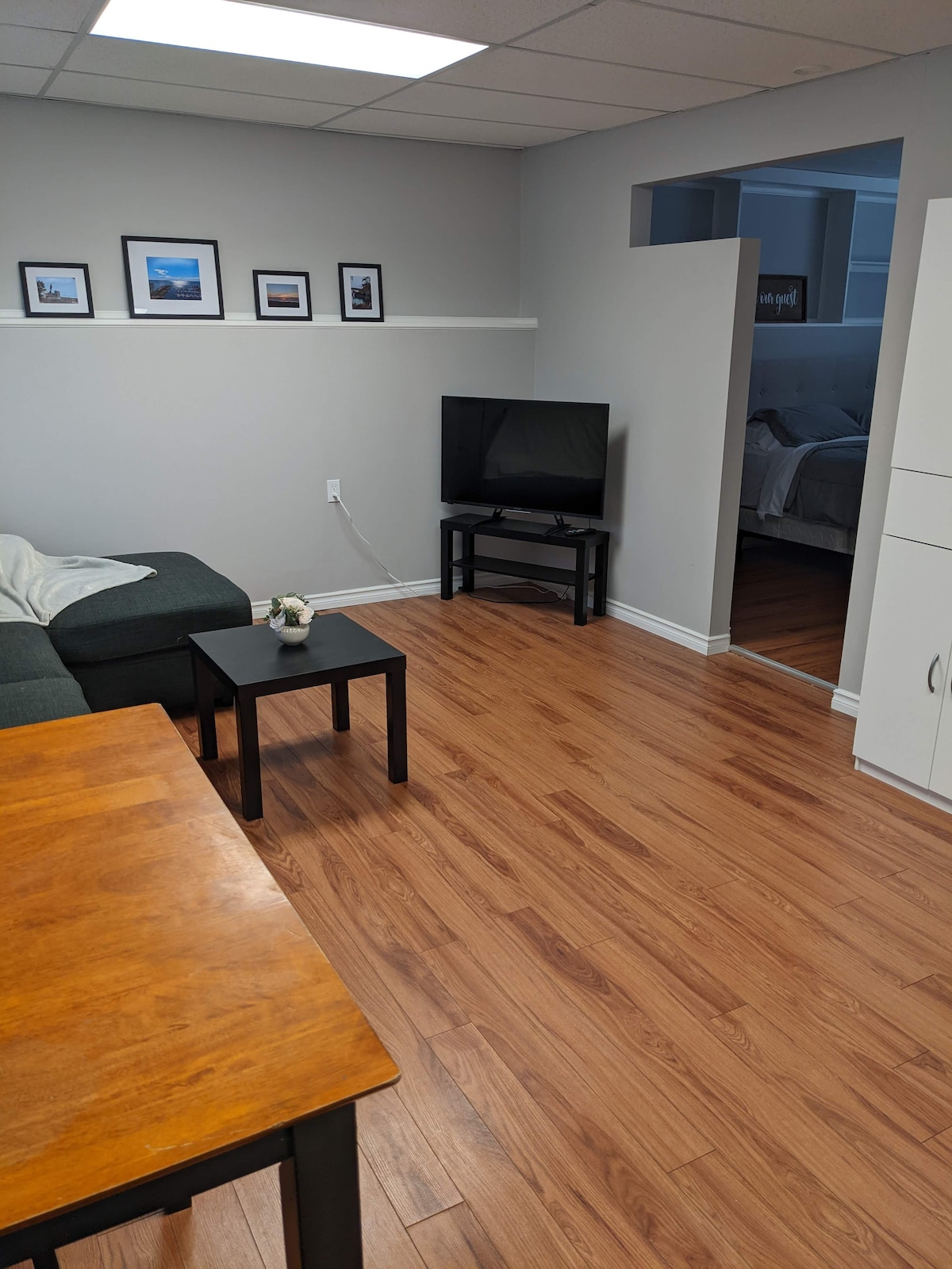 A comfortable living space is presented with a dark sofa and a small black table. A flat-screen television is positioned on a stand, and framed photographs adorn the wall. Light wood flooring complements the gray walls, adding to the room's welcoming atmosphere.