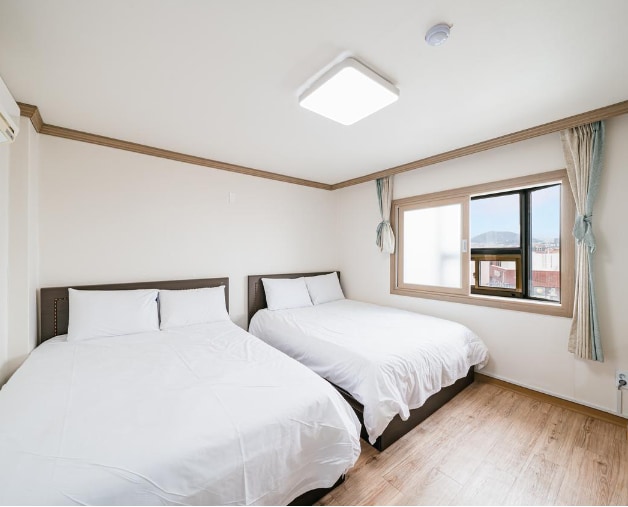 A bright and airy sleeping area features two spacious beds covered with white bedding. Natural light enters through a large window adorned with light curtains, offering a view of the outside. The wooden flooring complements the neutral-colored walls and ceiling.