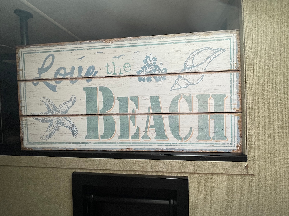A rustic wooden sign reading 'Love the Beach' is displayed prominently. The sign features beach-themed artwork, including a starfish and a shell, enhancing the coastal atmosphere within the space.