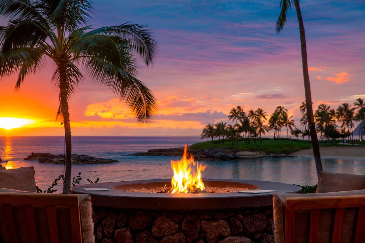 A fire pit is positioned in the foreground, surrounded by wooden lounge chairs. Vibrant hues of orange and purple fill the sky as the sun sets over the ocean. Palm trees frame the scene, highlighting the serene beachfront setting.