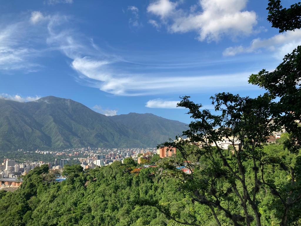 A panoramic view showcases lush green hills contrasted by the sprawling urban landscape below. The majestic mountains rise in the background under a clear blue sky with wispy clouds, providing a serene and tranquil setting.