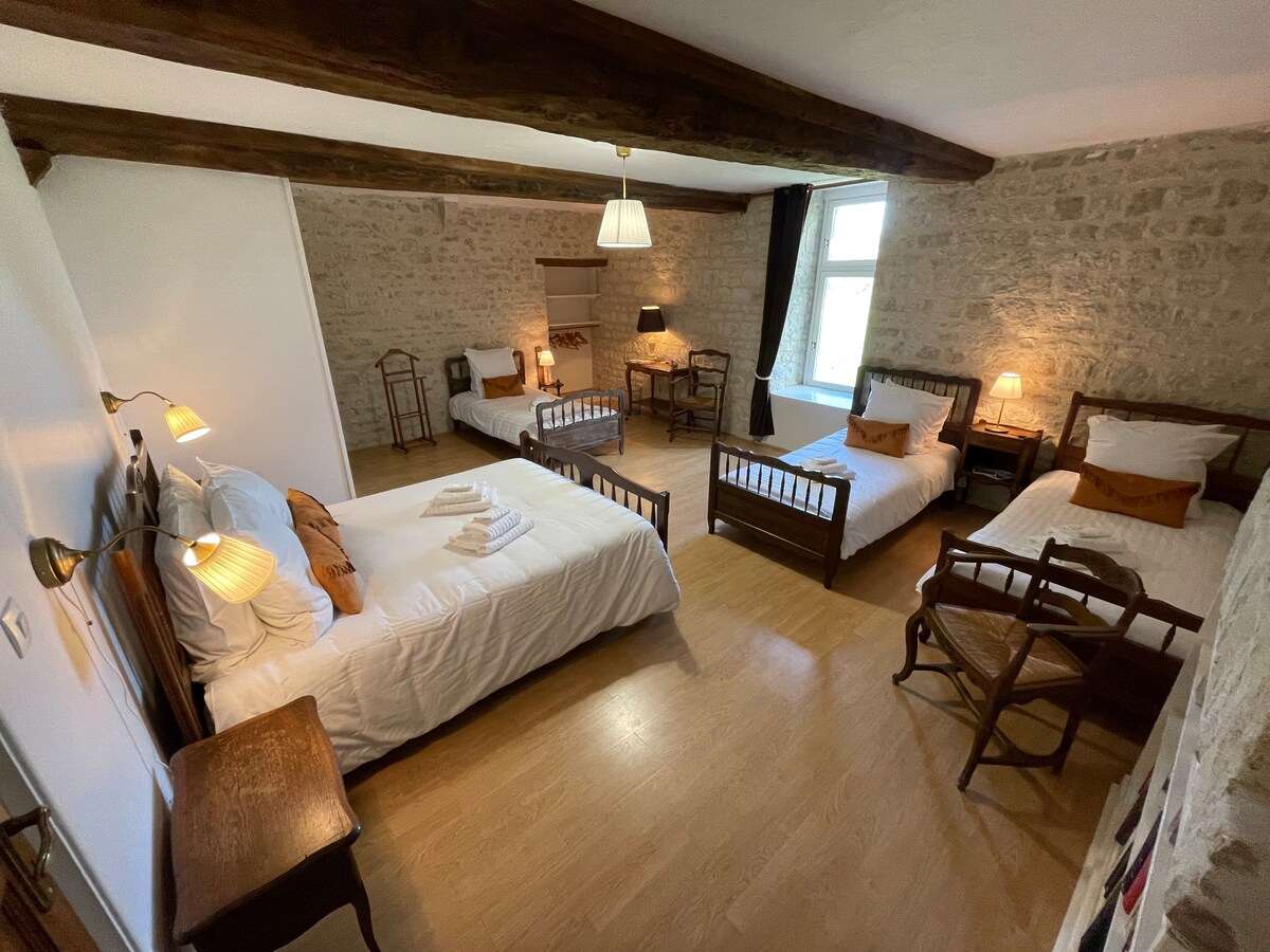 A spacious room features a double bed and three single beds arranged for comfort. Natural light filters through a large window, illuminating the rustic stone walls and wooden beams overhead. A small table and chairs add to the homely atmosphere.