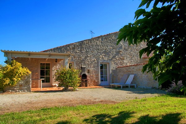 A spacious stone exterior is highlighted by a covered patio area, featuring a barbecue grill and two lounging chairs. Lush greenery surrounds the property, while bright blue skies provide a clear backdrop, creating an inviting outdoor setting for relaxation.