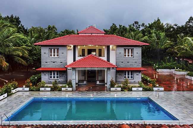 A spacious bungalow features a red-tiled roof and gray stone walls, framed by lush greenery. A rectangular swimming pool is centered in front, reflecting the surrounding area. The entrance is adorned with white pillars and flanked by neatly arranged flower beds.