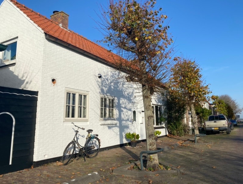 A charming white house with a red-tiled roof is displayed against a clear blue sky. A bicycle leans against the wall near the entrance, while trimmed trees frame the exterior. The paved area in front provides space for parking and walking.