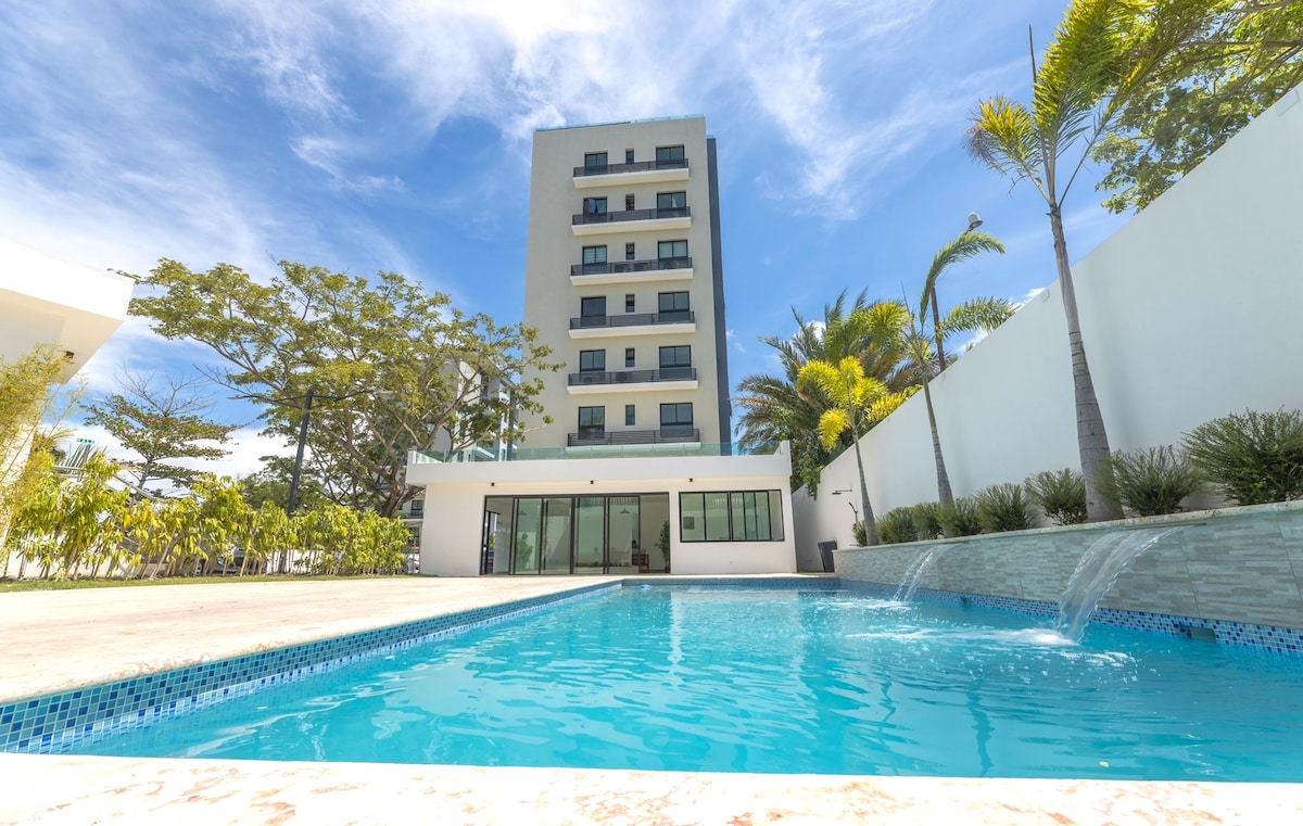 The modern building is seen beside a sparkling pool, featuring a serene outdoor space with palm trees and landscaped greenery. Sunlight reflects off the water, highlighting the inviting atmosphere of the area, perfect for relaxation.