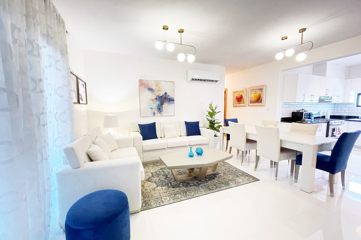 A bright living area features a comfortable white sofa accented with blue cushions, paired with a central coffee table. A decorative rug sits below, and a dining area with a light-colored table and chairs is visible in the background. Soft lighting is provided by stylish fixtures.
