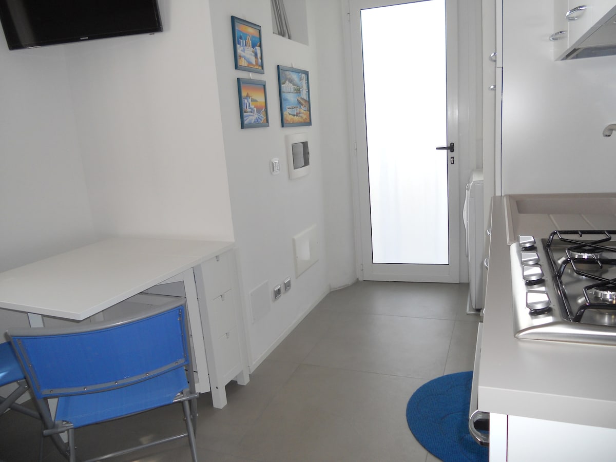 A small, well-lit kitchen area features a compact dining table and a blue chair, positioned next to a sleek stove. The wall displays three framed coastal artworks. A door leads to an exterior area, while a blue rug adds a touch of color to the tiled floor.