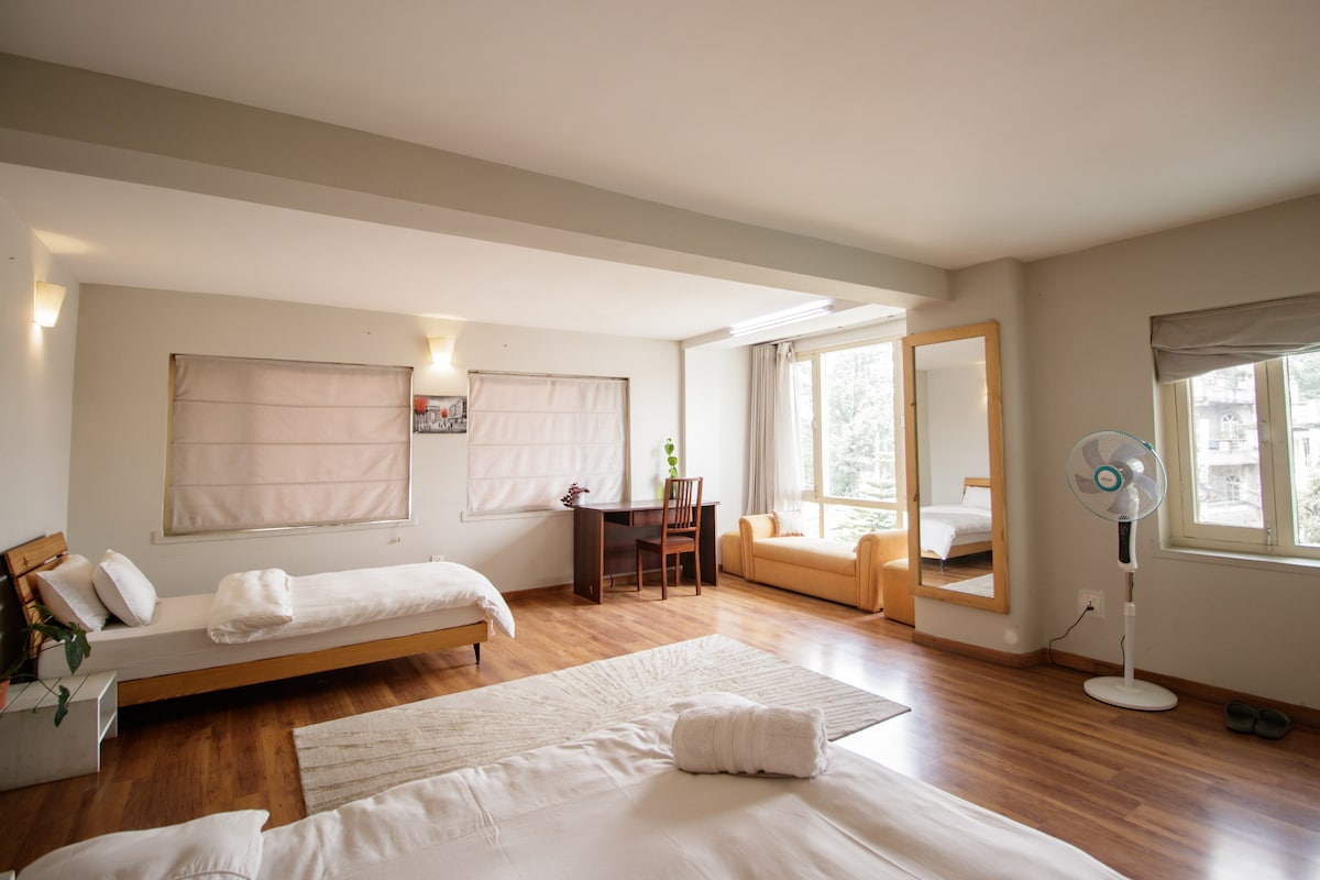 The studio apartment features light wooden flooring, with a spacious layout. It includes a cozy sitting area with a sofa, a work desk placed by a window, and beds arranged thoughtfully. Natural light enters through large windows, illuminating the inviting interior.