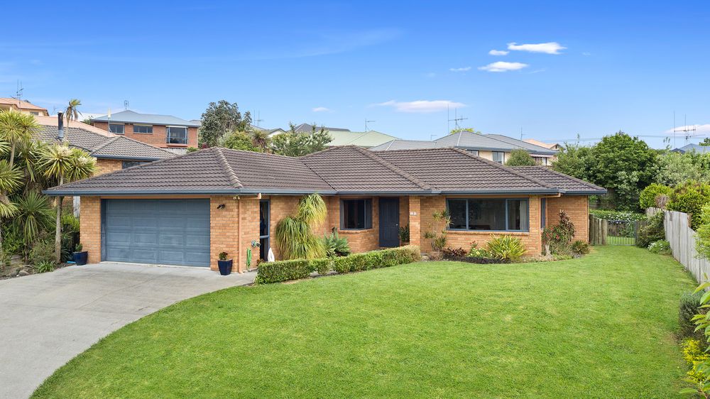 A single-story brick house is set on a well-maintained green lawn, flanked by various shrubs and plants. A concrete driveway leads to a garage on the left, while large windows offer views of the outdoor space. The sky above is clear and blue.