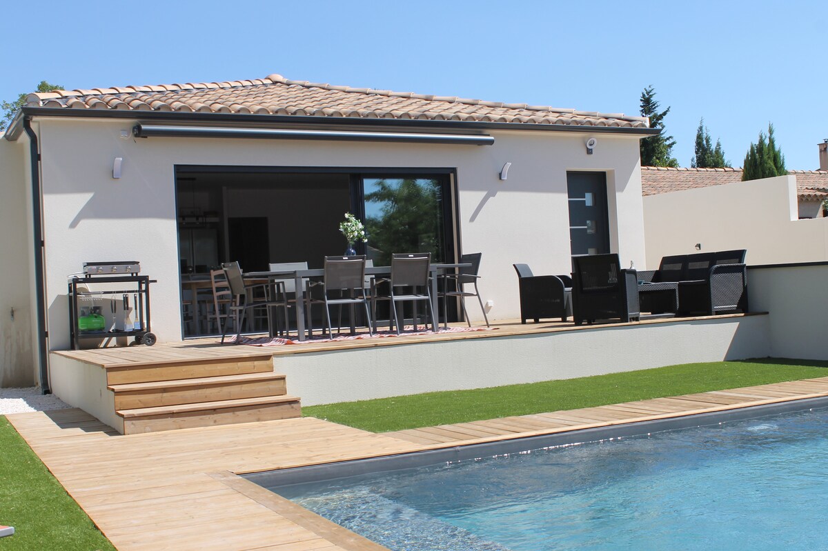 A modern villa features a spacious outdoor deck with seating and dining areas, surrounded by manicured grass. The swimming pool reflects clear blue skies, and large glass doors provide an open view into the interior living space.