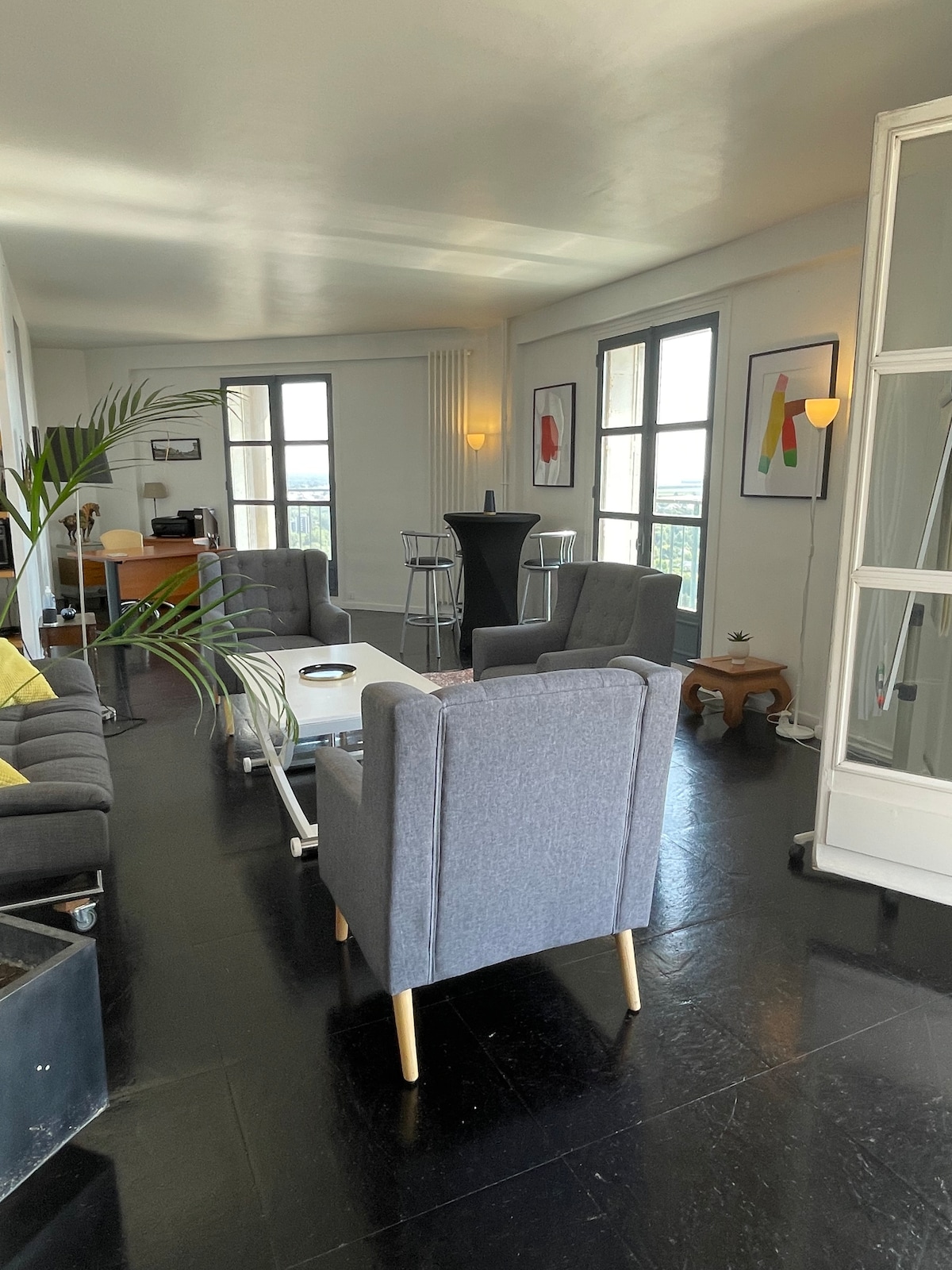 A spacious living area features two gray armchairs and a gray sofa, arranged around a low coffee table. Large windows provide natural light and stunning views of the surroundings. Art pieces adorn the walls, and a plant adds a touch of greenery to the space.