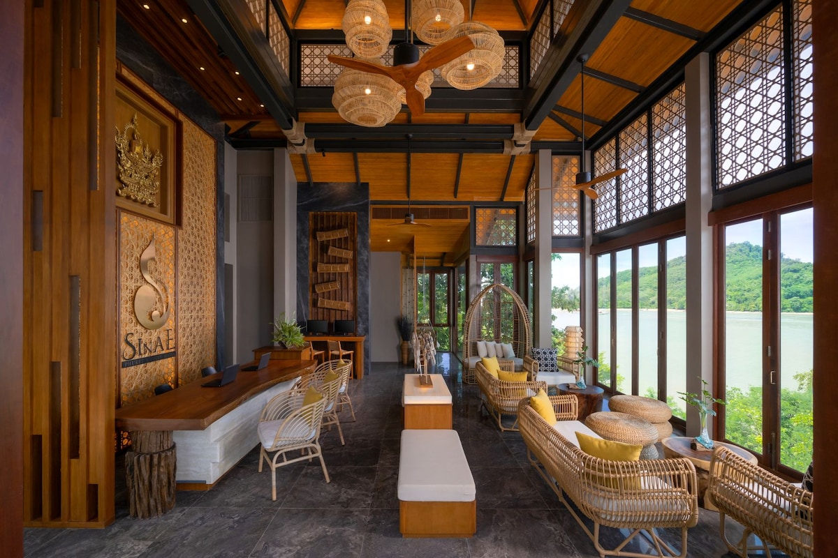 The lobby area features high ceilings with wooden beams and large glass windows, providing a view of the surrounding landscape. Natural decorations, including plants and wooden accents, complement the wicker seating arrangements. Elegant light fixtures hang overhead, enhancing the spacious and welcoming atmosphere.