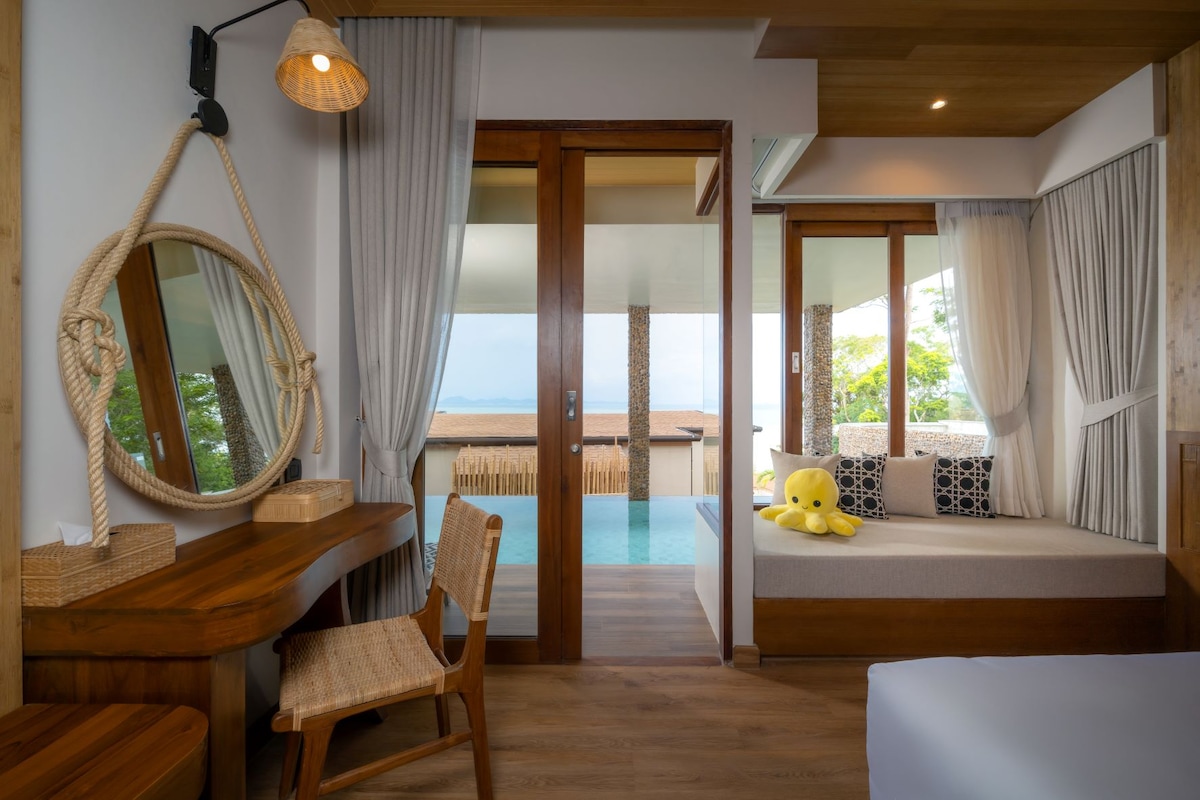 A cozy studio features a wooden interior with large glass doors opening to a private pool area. A comfortable seating nook with a plush cushion is positioned near the entrance. A stylish mirror and a wooden desk add functionality to the space.