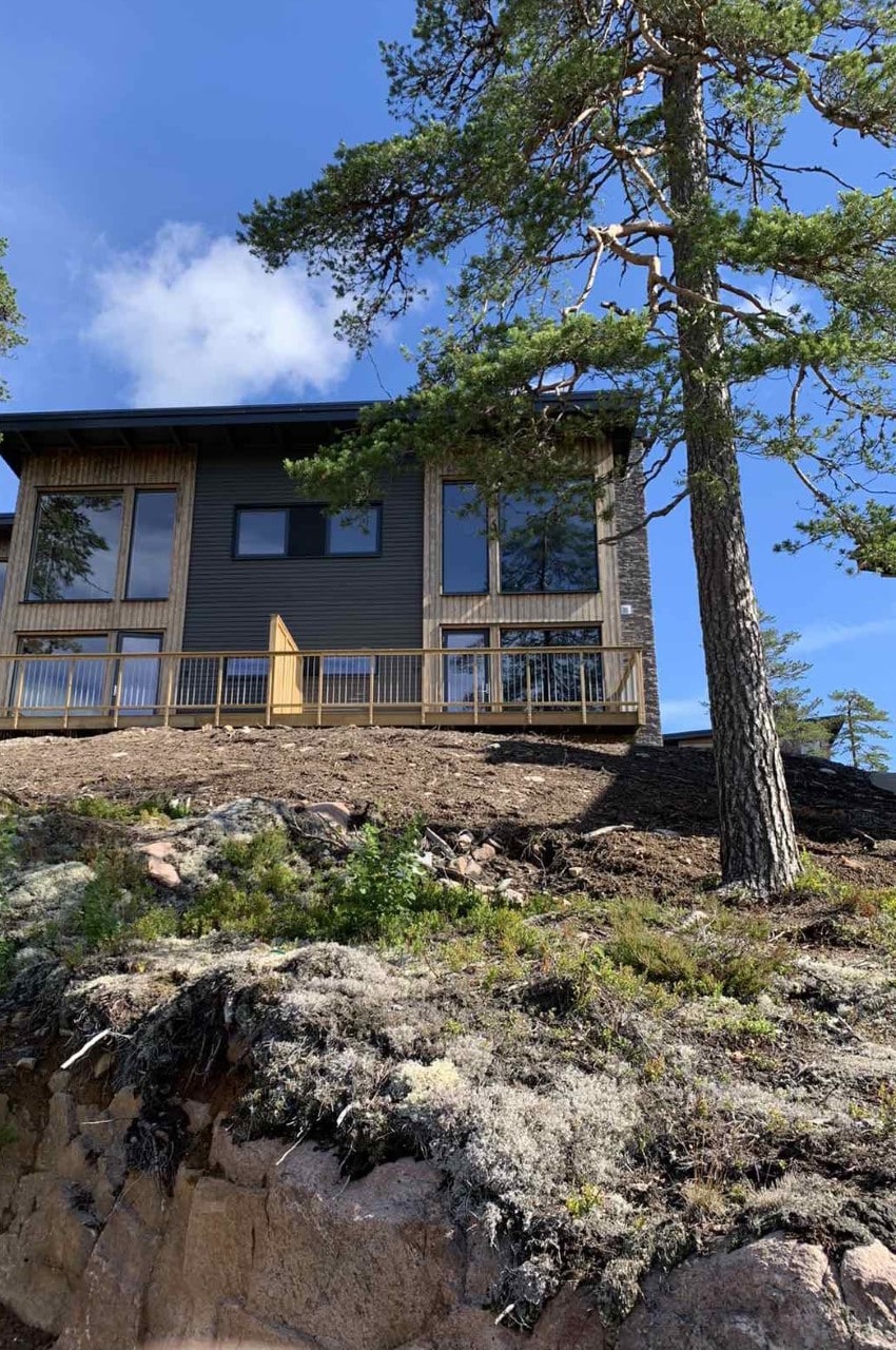 The two-story cabin is set against a backdrop of natural rock formations, featuring large windows that provide views of the surrounding landscape. A wooden deck extends from the structure, creating an inviting outdoor space. Tall pine trees frame the cabin, enhancing its mountain setting.