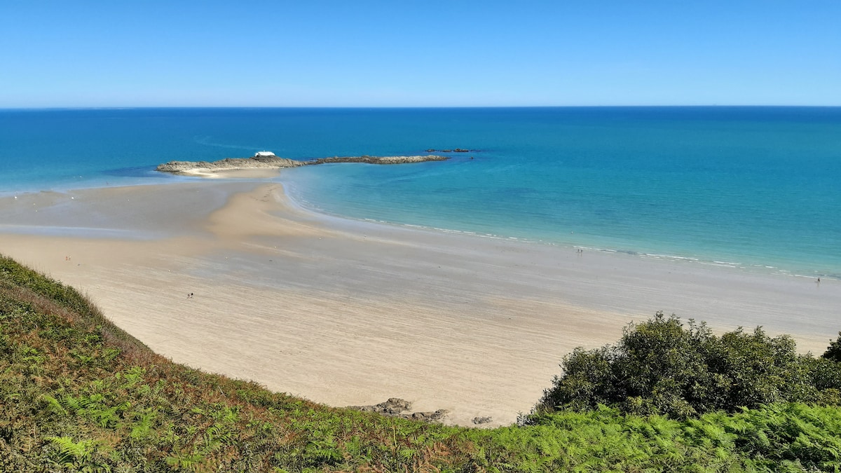 A serene beach scene is depicted with soft, sandy shores meeting calm turquoise waters. Gentle waves lap at the shoreline, while a rocky outcrop is visible in the distance, framed by lush greenery in the foreground, creating a peaceful coastal landscape.