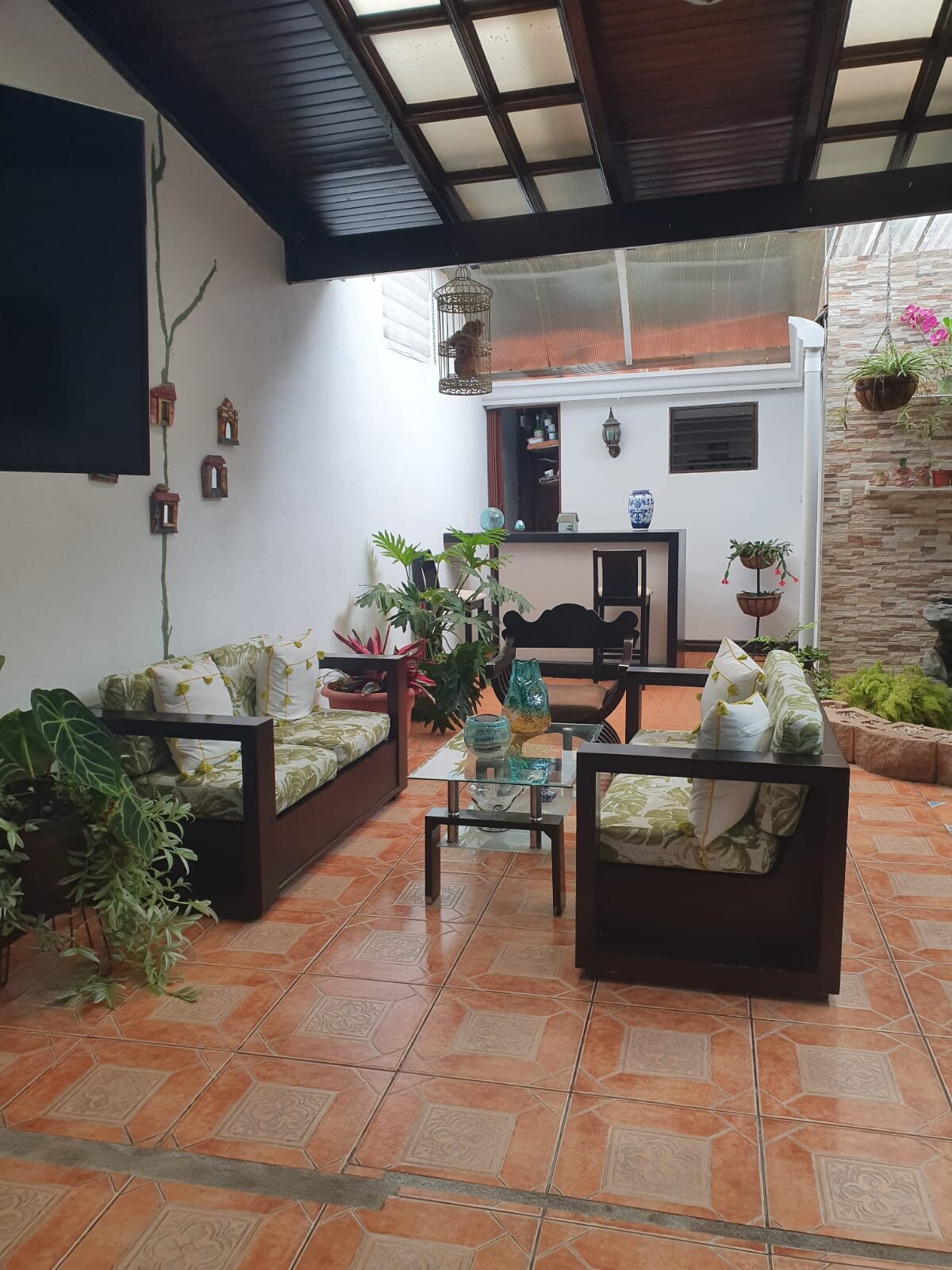 An inviting shared patio features two dark wooden seating areas with plush cushions, surrounded by lush greenery. A small glass table sits at the center, while decorative plants are arranged against the walls, creating a serene outdoor atmosphere.