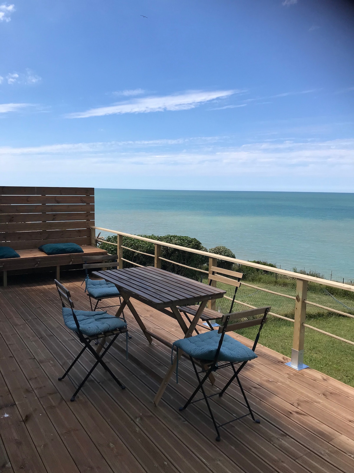 An outdoor terrace features a wooden deck with a dining table and four folding chairs. A lounge area is set with two cushioned benches facing the sea. The view includes a clear blue sky and the tranquil waters of the ocean.