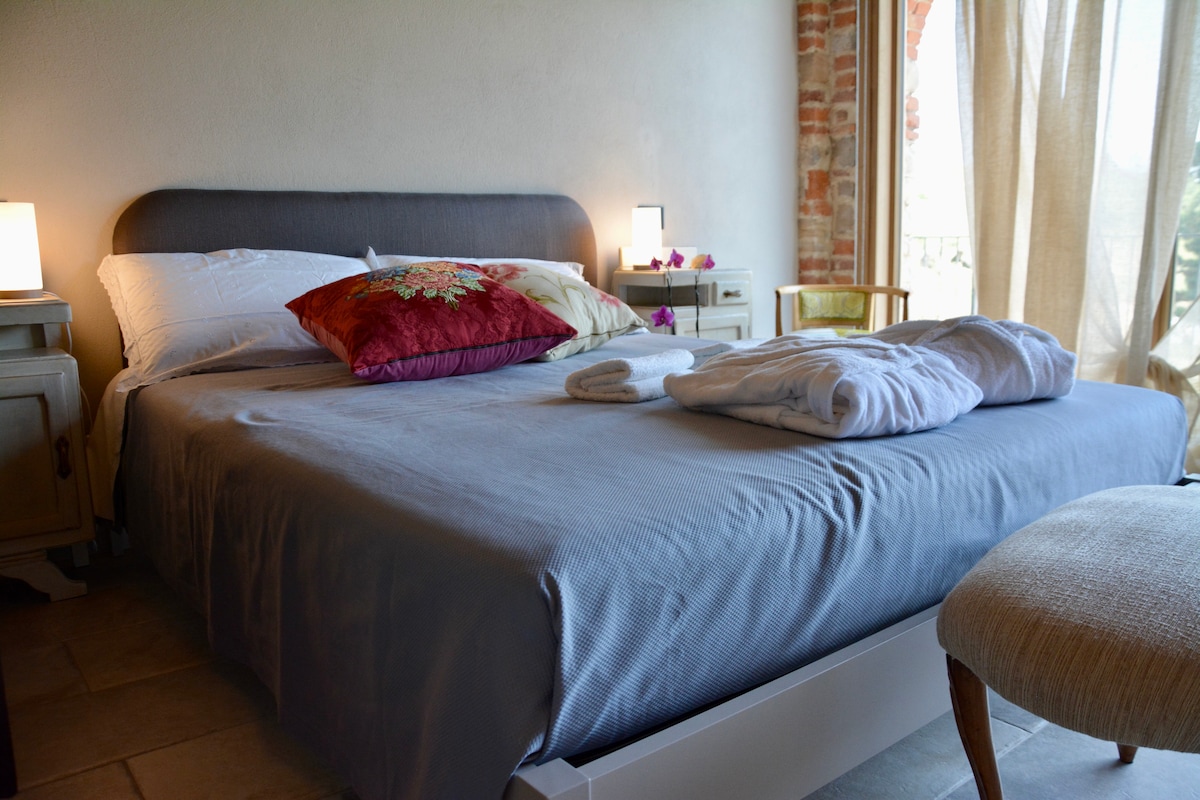 A spacious bed is dressed in soft linens, complemented by decorative pillows. Nearby, a small chair and softly glowing lamps are visible. Natural light filters through sheer curtains, accentuating the warm tones of the room's rustic elements.