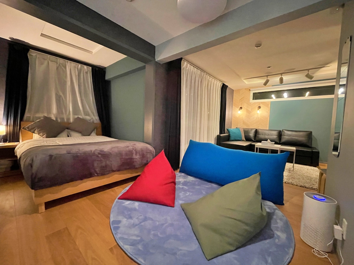 A spacious room features a double bed with soft bedding and two decorative pillows. A cozy seating area with a circular rug displays several assorted cushions. Large windows invite natural light, while dark curtains provide privacy.