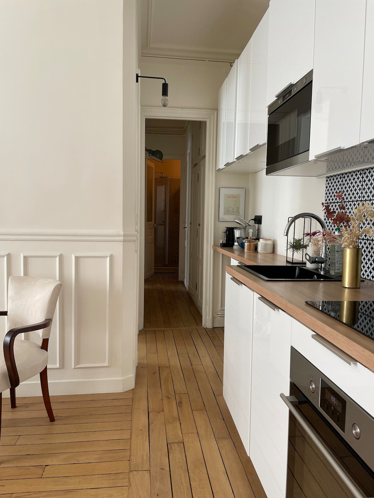 A modern kitchen area is visible, featuring sleek white cabinets and a wooden countertop. Kitchen appliances, including a coffee maker and oven, are positioned along the wall. The room is connected to a corridor leading to other areas of the apartment, with wooden flooring throughout.