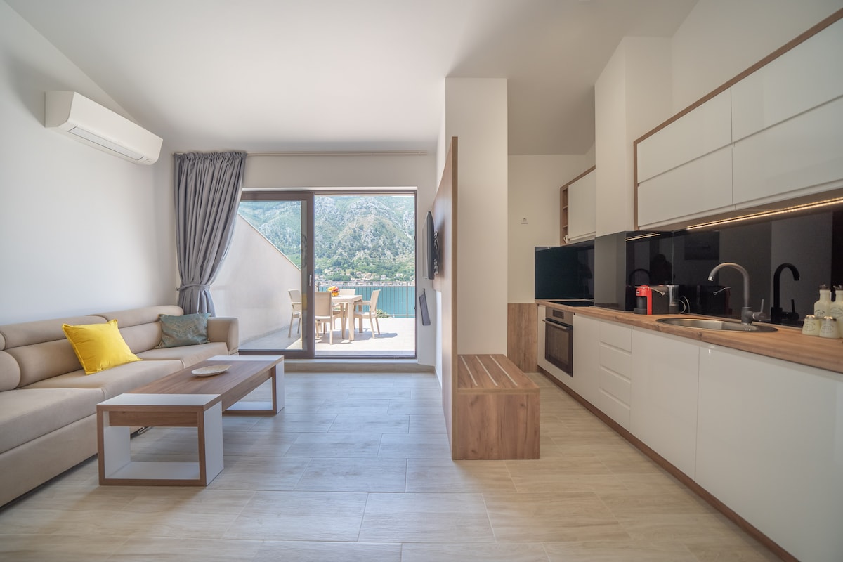 A bright living space features a comfortable sofa and a wooden coffee table, complemented by large glass doors leading to a balcony with views of the sea. Modern kitchen elements include sleek cabinetry and essential appliances, while natural light enhances the open layout.