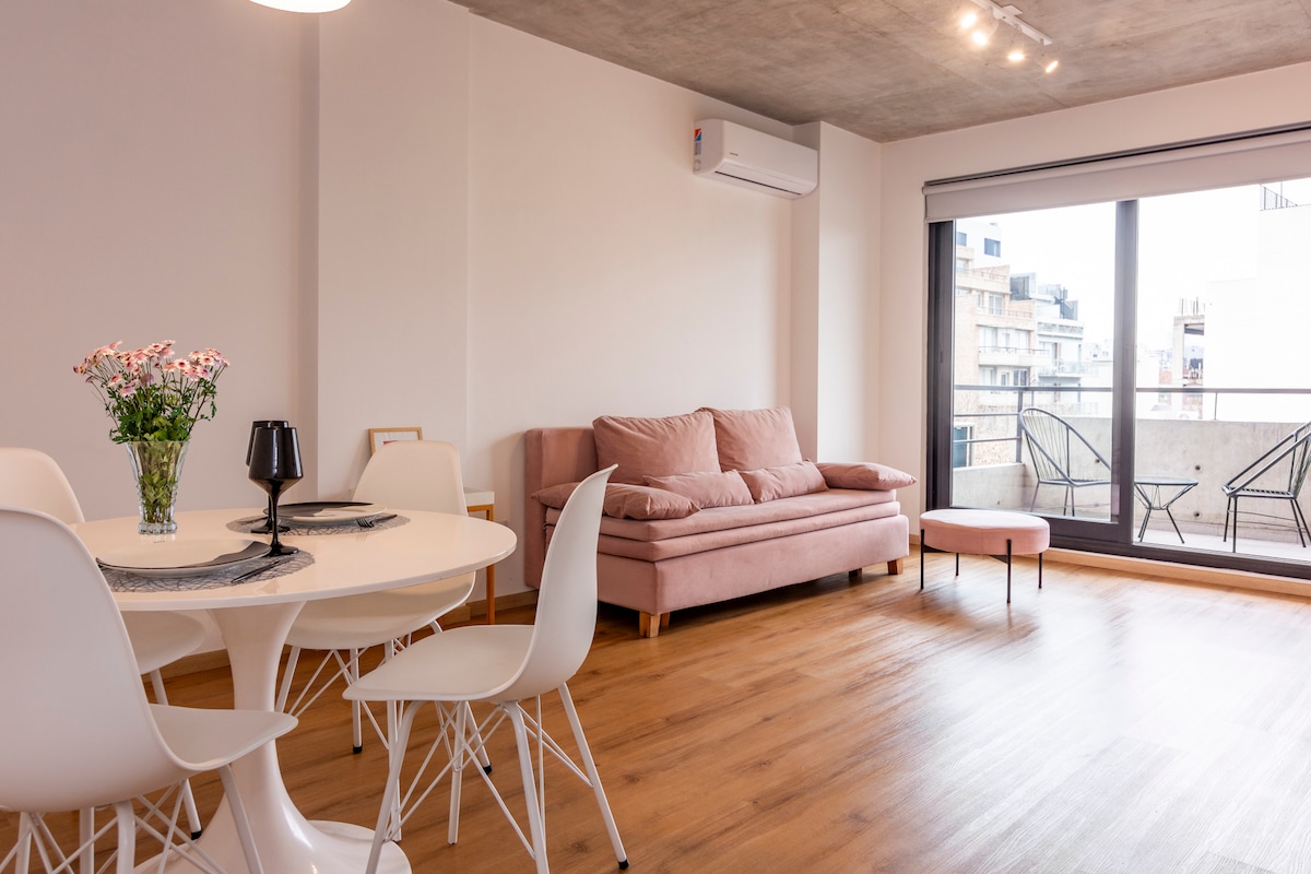 A light-filled living space features a cozy pink sofa and a round dining table surrounded by white chairs. A small side table complements the seating area. Floor-to-ceiling windows open to a balcony, with natural wood flooring enhancing the simple and modern design.