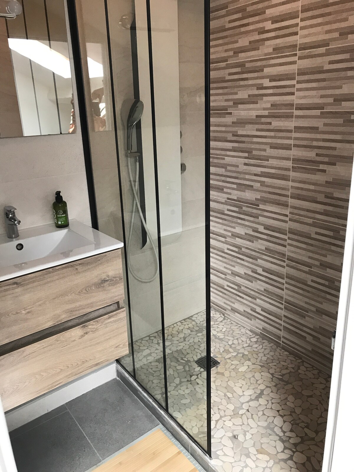 A modern bathroom features a glass-enclosed shower with a rain showerhead and a textured stone floor. A sleek vanity with wooden drawers provides storage, while a mirrored cabinet reflects natural light from the unique ceiling design.