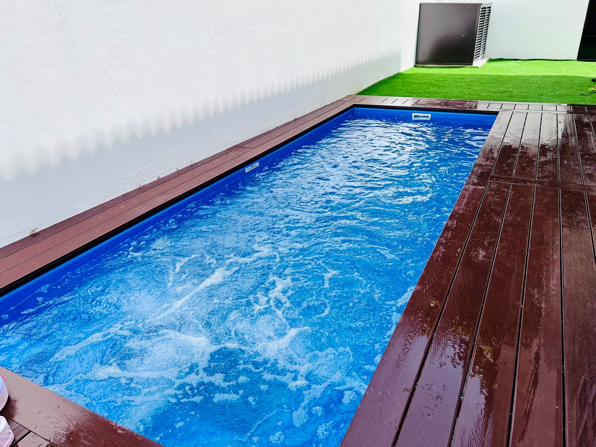 A rectangular swimming pool is set within a wooden deck, featuring clear blue water and gentle waves. The pool area is surrounded by greenery, offering a serene outdoor space for relaxation. A portion of the adjacent lawn is visible, enhancing the outdoor ambiance.