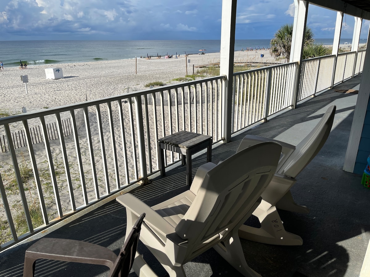 A spacious beachfront deck is equipped with two Adirondack chairs, providing direct views of the sandy shore and ocean waves. The railing allows for unobstructed sightlines to the beach, where various beachgoers can be seen enjoying the day.