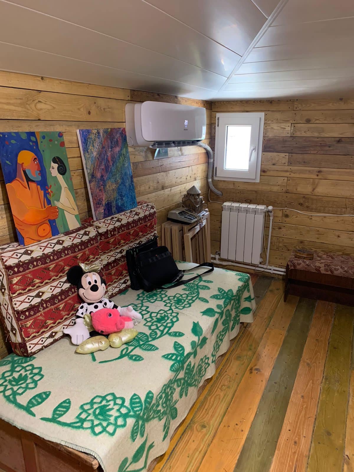 The attic room is adorned with wooden walls and features a small window offering natural light. A decorative green blanket is spread across two single beds, accompanied by a plush toy and a handbag. An air-conditioning unit provides comfort, while a wooden bench offers additional seating.
