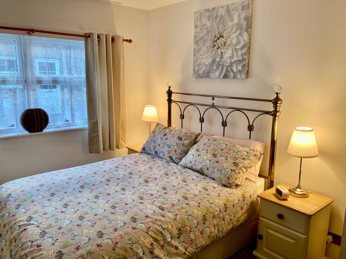 A cozy bedroom features a spacious bed adorned with floral-patterned bedding. Soft light emanates from two bedside lamps, illuminating the room. A piece of wall art hangs above the bed, with natural light filtering through sheer curtains on the window.