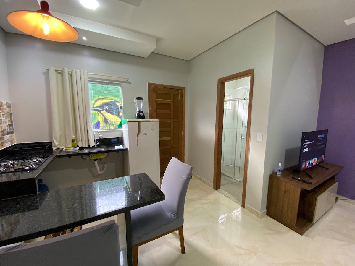 A modern kitchen area features a dark granite countertop, equipped with a blender and stovetop. Nearby, a small white dining table with two chairs is positioned. A door leads to a bright bathroom, while a television stands against the wall in the adjoining living space.