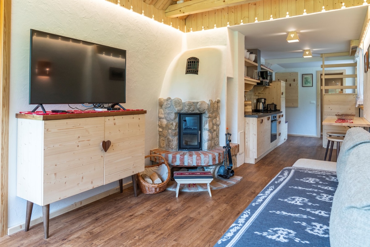 A welcoming living area is seen with a flat-screen TV mounted on a light wooden cabinet. A traditional stone fireplace adds charm, while a well-equipped kitchen is visible in the background. Soft lighting enhances the cozy ambiance, complemented by warm wooden accents throughout the space.