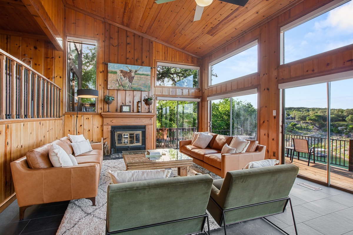 A spacious living room features wooden paneling and high ceilings, with large windows offering views of the surrounding landscape. Comfortable leather sofas and armchairs are arranged around a coffee table, while a fireplace adds a cozy touch to the space.