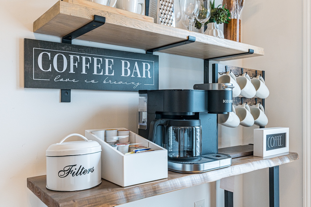 A coffee station is presented with a brewing machine on a wooden shelf. Above, a decorative sign reads 'COFFEE BAR.' A canister labeled 'Filters' and a box for coffee supplies are neatly arranged beside a row of cups hanging below the shelf.