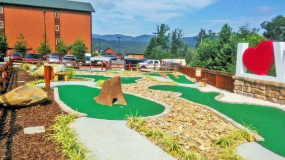 A mini-golf course is depicted with a vibrant green surface and a variety of stone features. Natural landscaping with small rocks and shrubs is evident throughout the course. Mountains can be seen in the background, along with parked cars and buildings nearby.