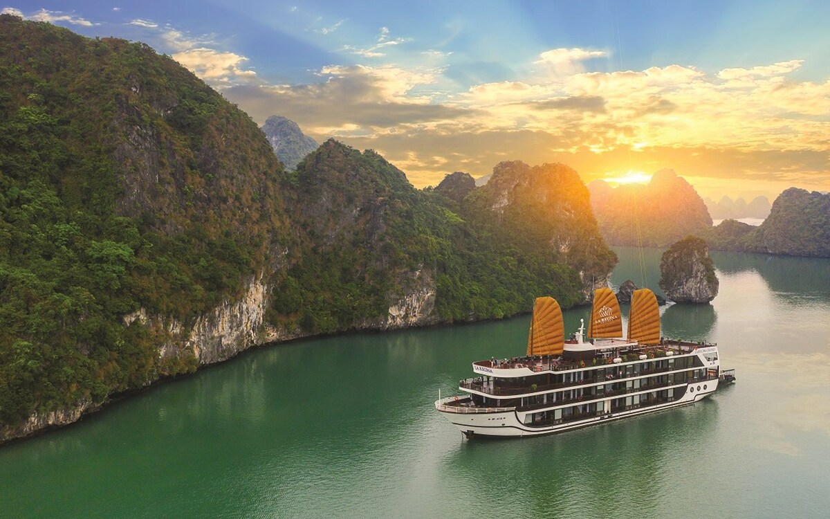 A spacious cruise ship is anchored in calm waters, framed by lush green mountains and rocky formations. The ship features distinctive orange sails, with a vibrant sunset casting warm tones across the tranquil bay.