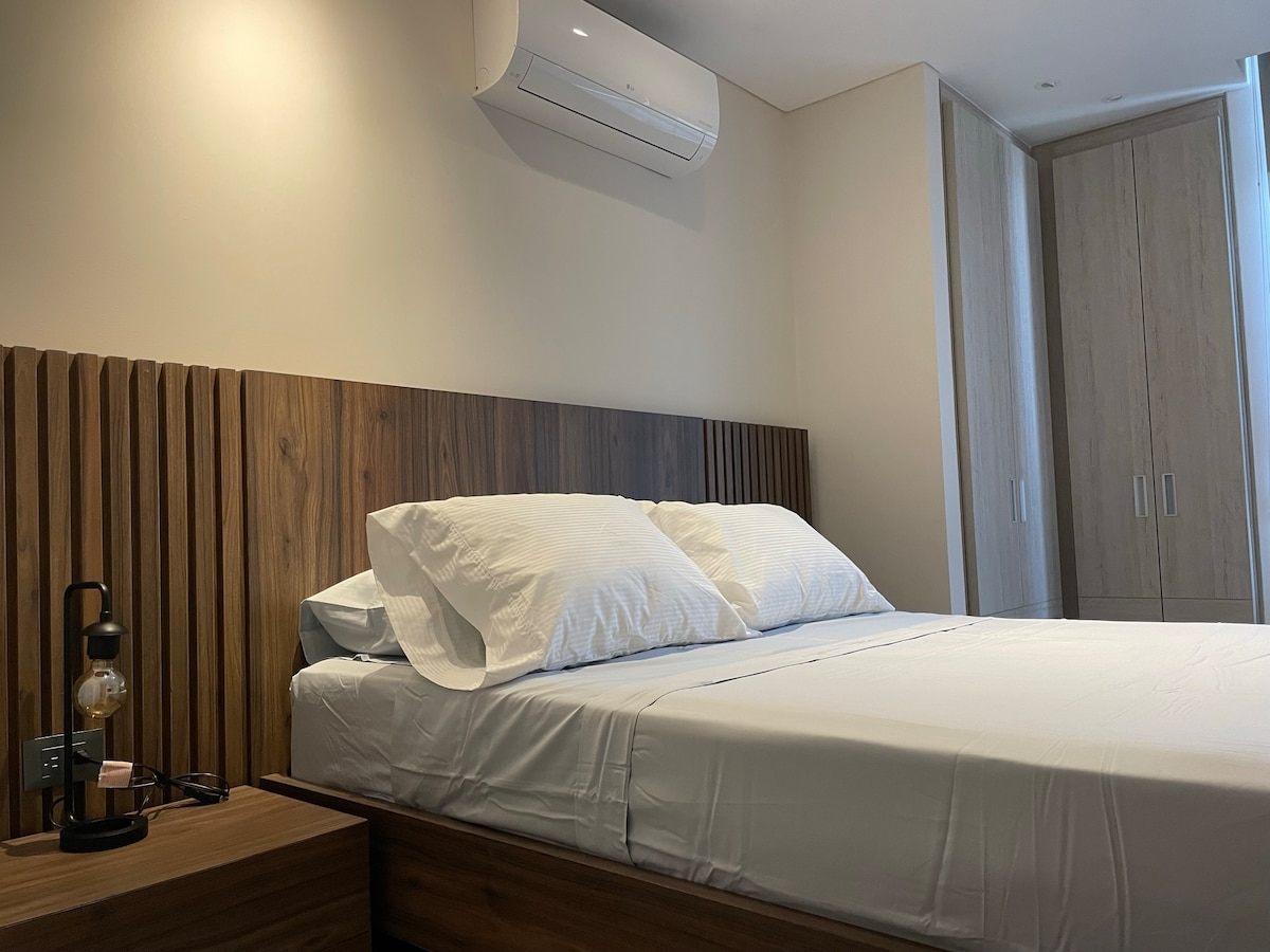 A spacious bedroom features a wooden bed frame with a large upholstered headboard. Two plump white pillows rest on the neatly made bed. An air conditioning unit is mounted on the wall, and a wardrobe stands in the background, offering ample storage space.