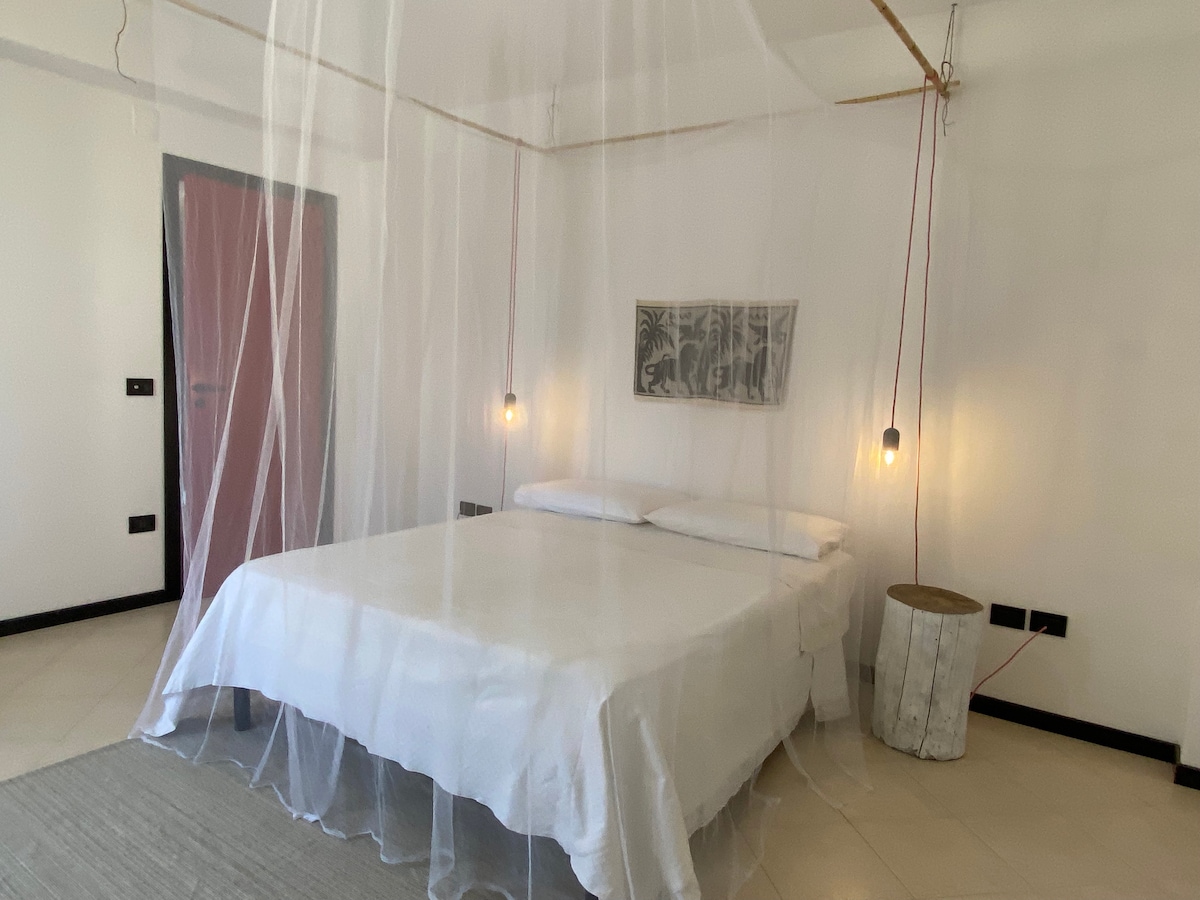 A serene bedroom features a spacious bed draped with a light sheer canopy. Soft lighting is provided by pendant lamps on either side, illuminating the minimalist decor. The walls are painted in a neutral tone, contributing to a calm atmosphere.