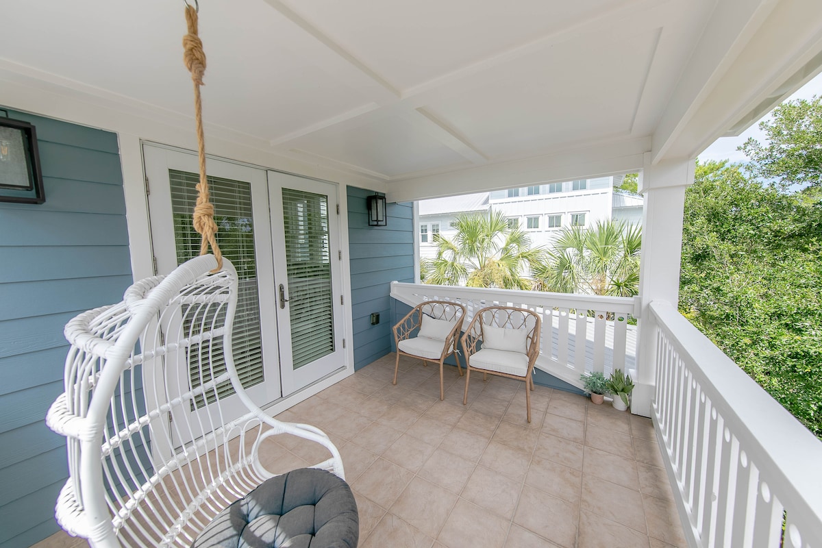 A spacious porch features a hanging chair alongside two woven chairs. Light-colored tile flooring complements the blue siding of the home. Large windows and doors allow natural light to fill the space, while lush greenery is visible in the background.