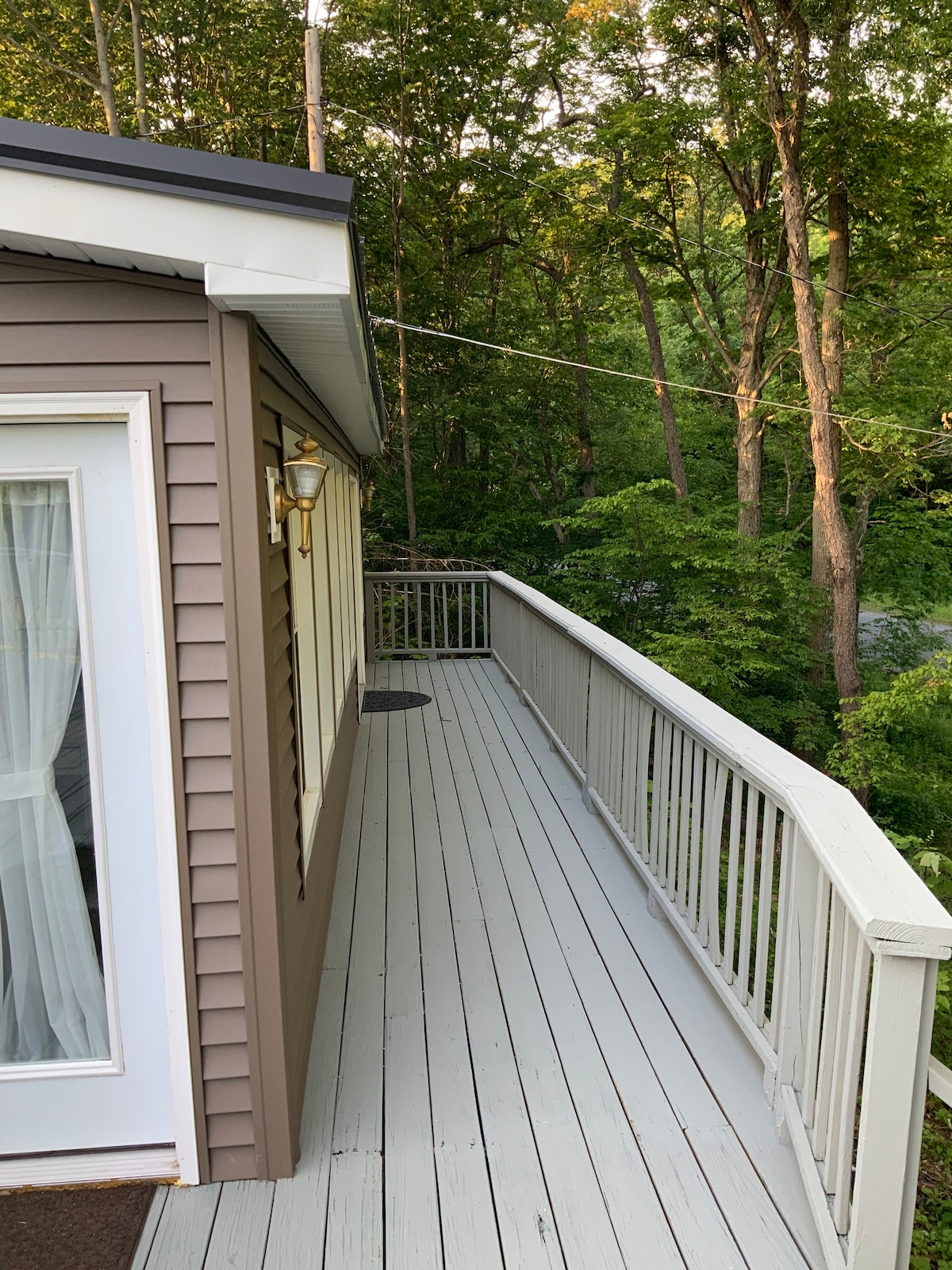 A wooden deck extends along the side of the house, bordered by a white railing. Lush greenery fills the view beyond, contributing to a serene and natural setting. Soft morning light reflects off the surfaces, creating a calm atmosphere.
